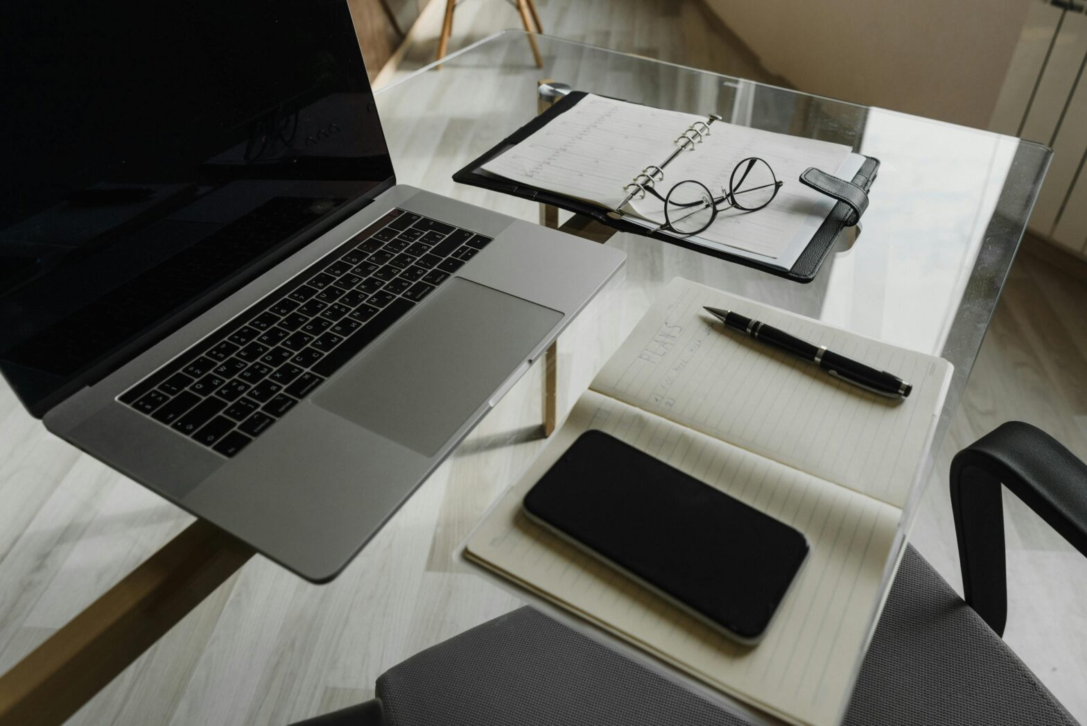 An image of a glass table top. Placed on top include a macbook, an iphone, mutlple note pads and glasses